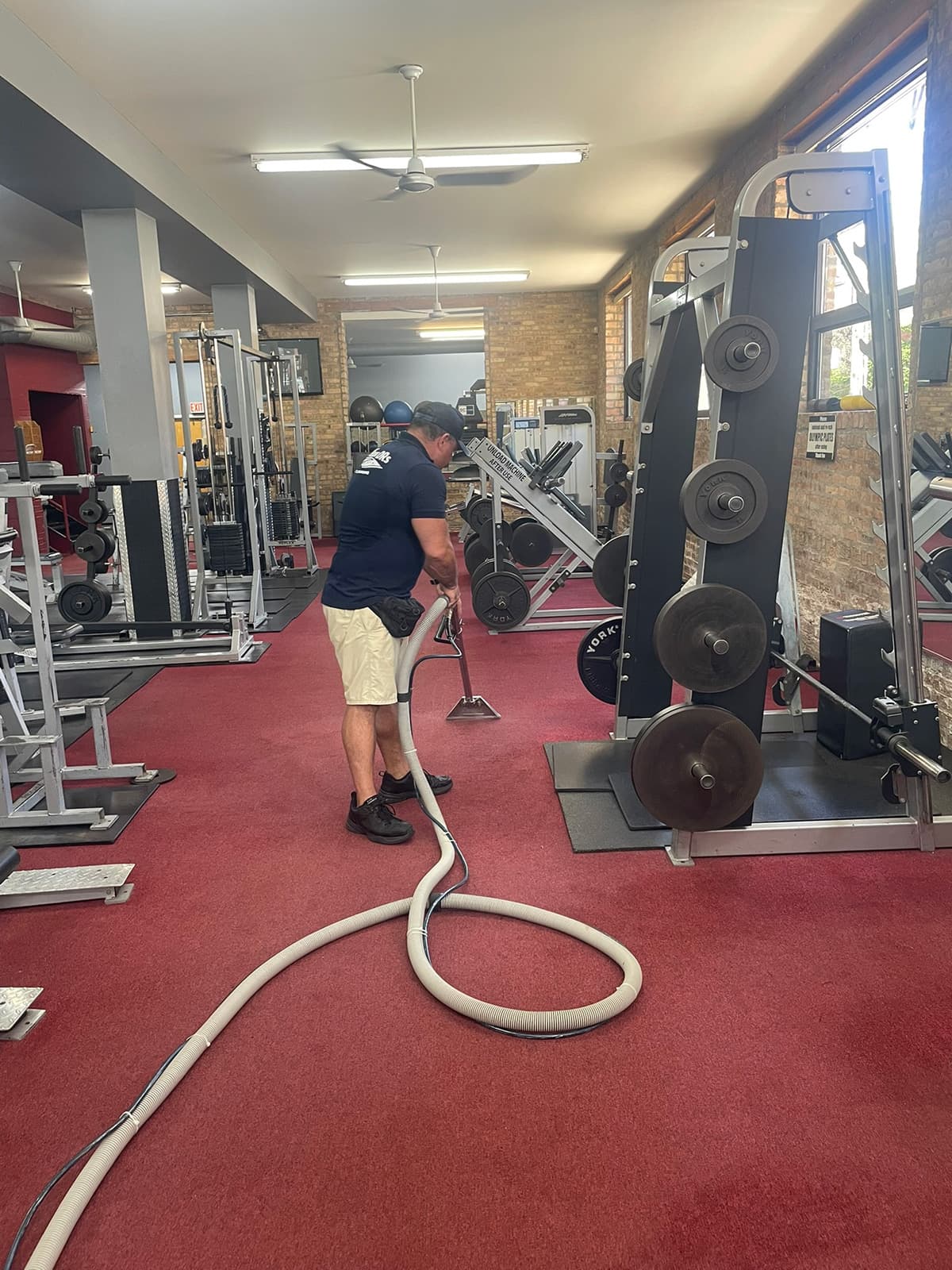 A technician extracting red carpet on a gym floor between weight racks