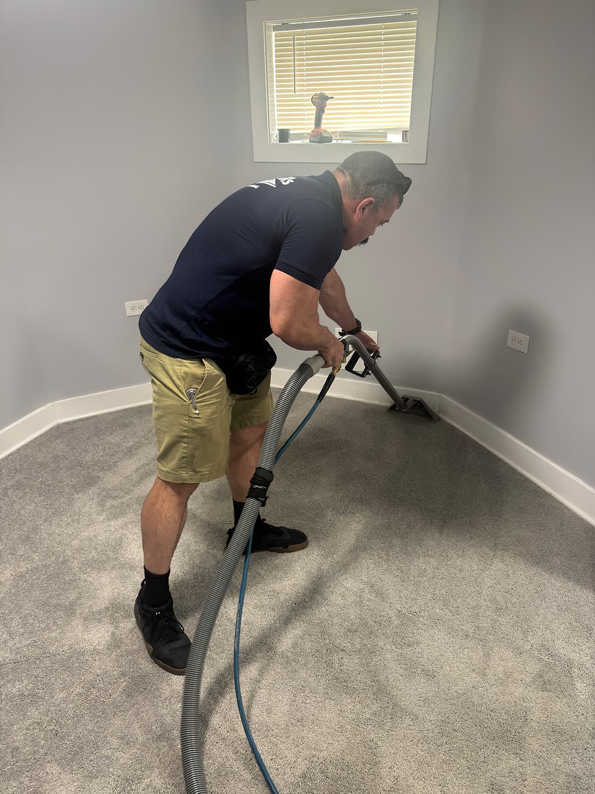 A technician in a branded shirt drawing an extraction wand along the baseboard of a grey bedroom carpet